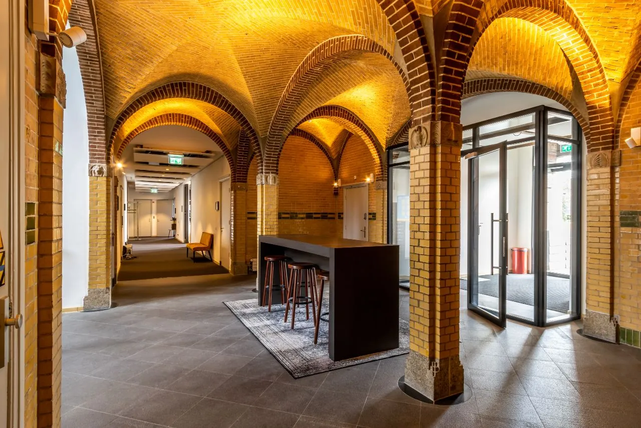 Entreehal van een karakteristiek gebouw aan de Lage Naarderweg met bakstenen gewelven, een hoge tafel en moderne inrichting.